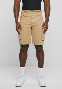 Shorts cargo en khaki en coton, avec des poches latérales, une coupe régulière et une fermeture à bouton. Portés avec un débardeur noir et des baskets blanches.