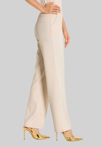Vrouw in beige hoge taille wijde pijpen broek en metallic gouden hoge hakken met spitse neus, staand opzij met hand op de heup.