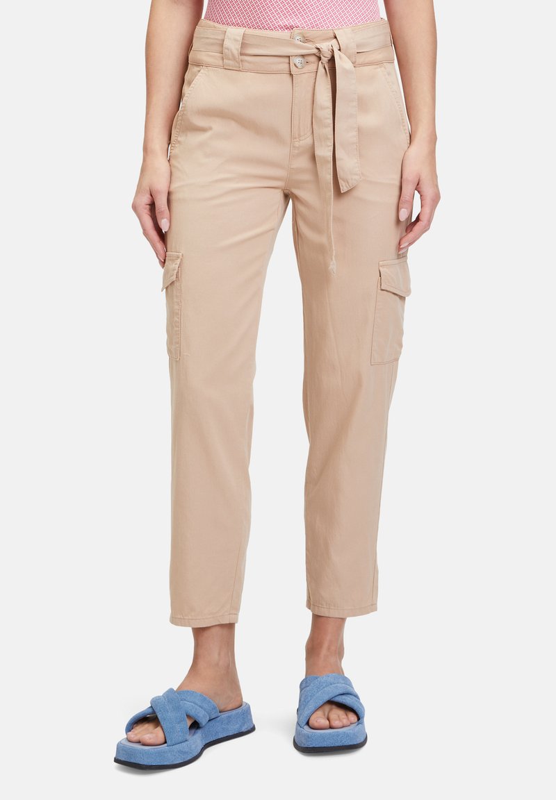Beige Cargo-Hosen mit einem Gürtel in der Taille, tapered Beinen und mehreren Taschen. Getragen mit blauen Sandalen mit Kreuzriemen. Der Stoff wirkt glatt und leicht.
