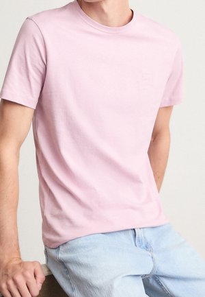 Mężczyzna noszący jasnoróżowy T-shirt z krótkim rękawem oraz jasnoniebieskie dżinsy, siedzący z jedną ręką spoczywającą na kolanie na jednolitym tle.