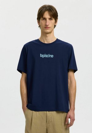Muž v tmavomodrom tričku so svetlomodrým nápisom „lapiscine“, béžové nohavice, stojaci pred jednofarebným svetlým pozadím.