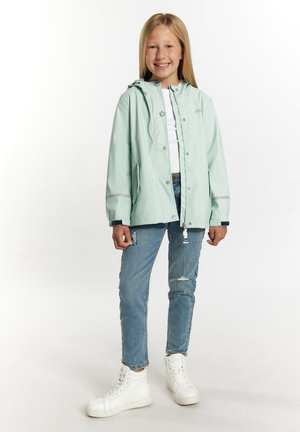 Lichtgroene waterdichte regenjas met een capuchon, zilveren accenten en drukknoopsluitingen, gecombineerd met blauwe ripped jeans en witte hoge sneakers.