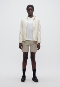 Νεαρό άτομο όρθιο, φορώντας λευκό τζάκετ με φερμουάρ, λευκό t-shirt, μπεζ σορτς, μαύρες κάλτσες και μαύρα αθλητικά παπούτσια, μπροστά σε απλό φόντο.