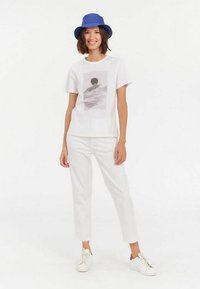 T-shirt blanche avec un imprimé graphique, associée à un pantalon blanc. Un chapeau seau bleu et des baskets blanches complètent le look décontracté.