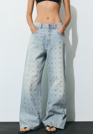 Svetlomodré široké rifle s kovovými štvorcovými cvokmi, nosené s rukami v vreckách a čiernymi žabkami, zobrazené od pása až po chodidlá.