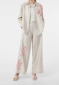 Beige tweedelig outfit met een knoopshirt met rode bladbroderie en wijde pijpen broek, gecombineerd met witte sandalen met bandjes.