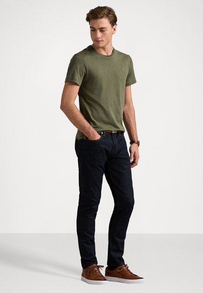 Olivnozelena bombažna majica, prilegajoča zasnova, kombinirana s temnimi jeans hlačami in rjavimi športnimi čevlji, z enostavnim logotipom na prsih.