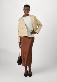 Giacca beige doppiopetto, top verde chiaro, lunga gonna in satin marrone, borsa nera, e scarpe basse stringate nere. Texture morbide in tutto.