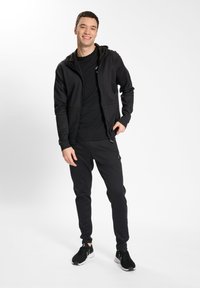 Outfit sportiv negru format dintr-o hanorac cu fermoar și pantaloni jogger taper, realizat dintr-un material neted cu o branding minim și un design ajustat.