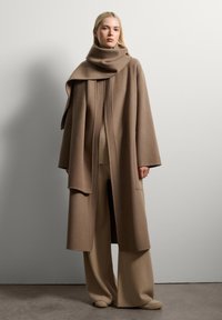 Lungo cappotto di lana beige con colletto alto e design drappeggiato. Abbinato a pantaloni larghi coordinati e calzature beige morbide.