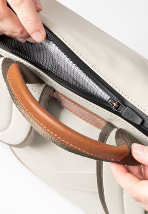 Hands opening a beige bag's black zipper near a brown padded handle, showing light-colored fabric interior with a grid pattern.