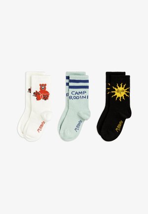 Tres pares de calcetines: blancos con un gráfico de oso rojo, verde menta con rayas marinas y texto, y negros con un gráfico de sol amarillo.