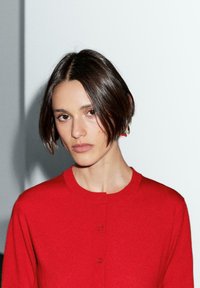 Červený pletený cardigan s kulatým výstřihem a zapínáním na knoflíky, s hladkou texturou a přiléhavým střihem. Jednoduchý design, bez vzorů.