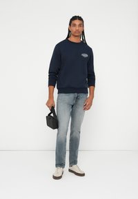 Marine crewneck genser med en liten hvit grafikk, kombinert med lyseblå jeans og beige sneakers. Holder en liten svart veske.