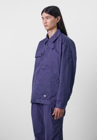 Lilla lett jakke med knappelukking på kragen, frontlomme og brystlomme med lapp. Stoffet ser glatt ut med minimal tekstur.