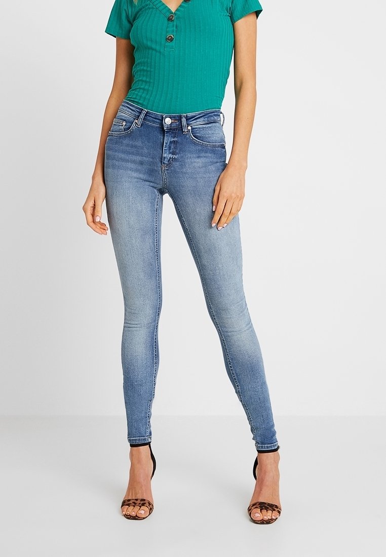 Jean en denim bleu taille haute avec une coupe slim, finition délavée, cinq poches et coutures contrastées, associée à un haut côtelé vert.