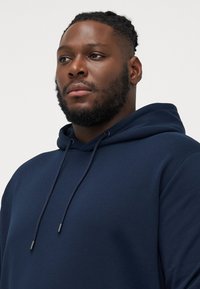 Marineblauwe hoodie met trekkoord, gemaakt van textuurstof. Heeft een vooraanstaande kangoeroezak en een ontspannen pasvorm.