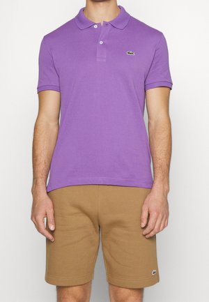 Mann trägt ein lila kurzärmeliges Poloshirt und beige knielange Shorts, steht vor einem einfachen weißen Hintergrund.