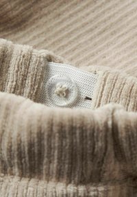 Tissu beige avec des plis de stries texturées recouvrant un textile blanc avec un bouton clair et un détail de fil beige. Gros plan de la zone de bouton.