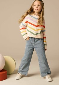 Pull en tricot multicolore avec un motif en chevrons, associé à un pantalon bleu à larges rayures. Le modèle porte des baskets blanches. Fond aux tons neutres.