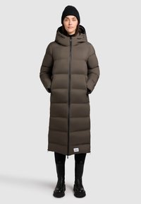 Vrouw in een lange bruine pufferjas met capuchon, zwarte beanie, zwarte legging en zwarte laarzen, staande voor een effen lichte achtergrond.