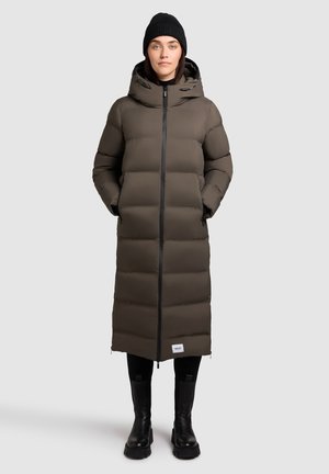 Vrouw in een lange bruine pufferjas met capuchon, zwarte beanie, zwarte legging en zwarte laarzen, staande voor een effen lichte achtergrond.
