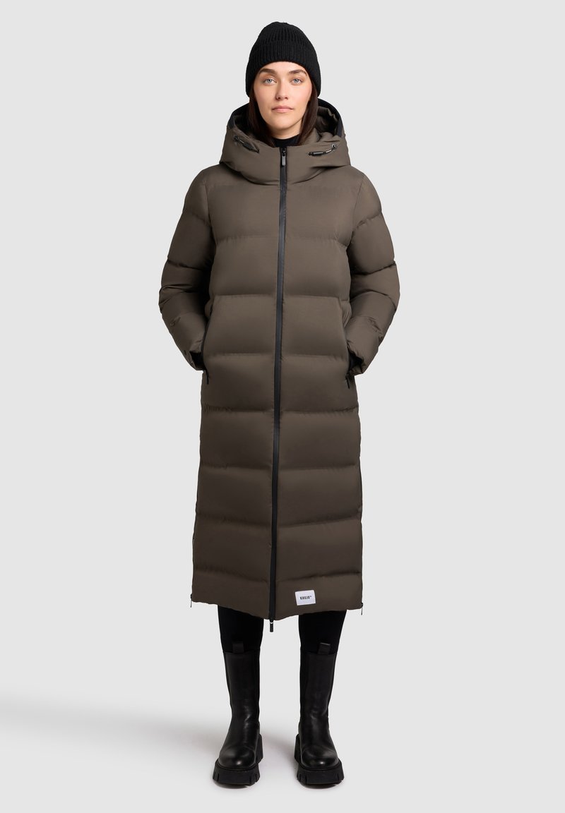 Vrouw in een lange bruine pufferjas met capuchon, zwarte beanie, zwarte legging en zwarte laarzen, staande voor een effen lichte achtergrond.