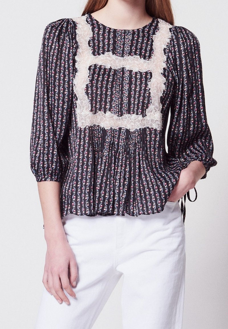 Femme portant un chemisier noir à motifs floraux avec des détails en dentelle blanche carrés, manches trois-quarts bouffantes, et un pantalon blanc, main dans la poche.