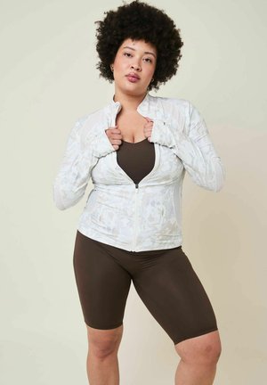Donna con capelli ricci che indossa una giacca bianca con zip sopra una canotta marrone e pantaloncini da bici marroni, in piedi davanti a uno sfondo semplice e chiaro.