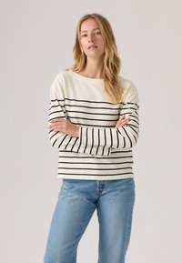 Weißes Langarmoberteil mit schwarzen horizontalen Streifen, runder Ausschnitt und geradem Schnitt; kombiniert mit hellblauen Denim-Jeans.
