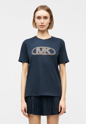 Γυναίκα που φοράει μπλε σκουρό μπλουζάκι με χρυσό διακοσμητικό λογότυπο "MK" και μια αντίστοιχη plisέ φούστα, στέκεται μπροστά σε απλό λευκό φόντο.