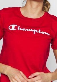 Rød bomulds-t-shirt med et hvidt "Champion" logo i kursiv. Klassisk rund hals og korte ærmer, glat tekstur og afslappet pasform.