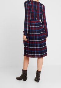 Kobieta w granatowej, czerwonej i białej w kratę sukience z długim rękawem i plisowanym dołem oraz ciemnobrązowych botkach za kostkę na białym tle.