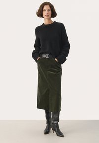 Schwarzer Strickpulli, grüne Cordrock und knielange schwarze Lederstiefel. Das Modell steht mit den Händen in den Rocktaschen und trägt einen schwarzen Gürtel.