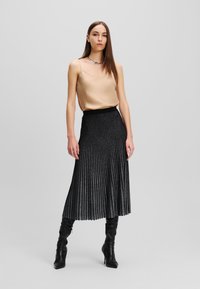 Beige satijnen camisole-top gecombineerd met een zwarte geplooide glinsterende rok. Het model draagt lange zwarte, aansluitende laarzen met een lichte glans op de stof.