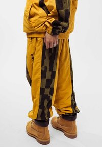 Yellow tracksuit featuring a textured dark brown abstract pattern on the sides, paired with tan boots. Material appears lightweight and durable.