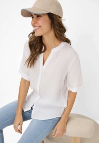 Letvægts hvid linnedskjorte med V-hals, korte opbundne ærmer og knaplukning, parret med blå stribede jeans og en beige kasket.