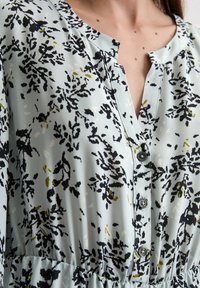Blumengemustertes Kleid aus hellblauem Stoff. Verfügt über einen knopfverschlossenen Ausschnitt und Akzentfarben in Schwarz und Olivgrün. Glatte Textur.