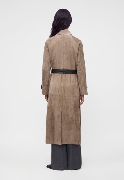 Femme portant un long manteau en suède beige avec une ceinture noire, un pantalon large sombre et des chaussures noires, debout de dos sur un fond uni.