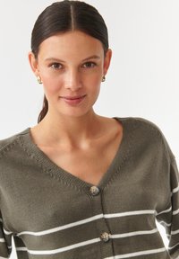 Olivgrüner Cardigan mit weißen Streifen, V-Ausschnitt und zwei großen, runden Knöpfen. Der Stoff wirkt weich mit einer Strickstruktur.