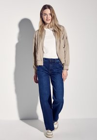 Béžová bomber bunda přes bílou košili, v kombinaci s tmavě modrými širokými džíny a bílými teniskami s béžovými detaily. Měkká struktura, ležérní styl.