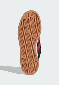 Suola di una scarpa con superficie in gomma strutturata a motivo ondulato, di colore marrone con tomaia nera e dettagli rossi.