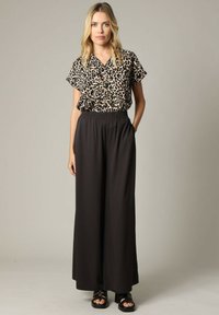 Korteblauwe blouse met luipaardpatroon, gedragen met zwarte wijde broek met elastische tailleband en platte zwarte sandalen.