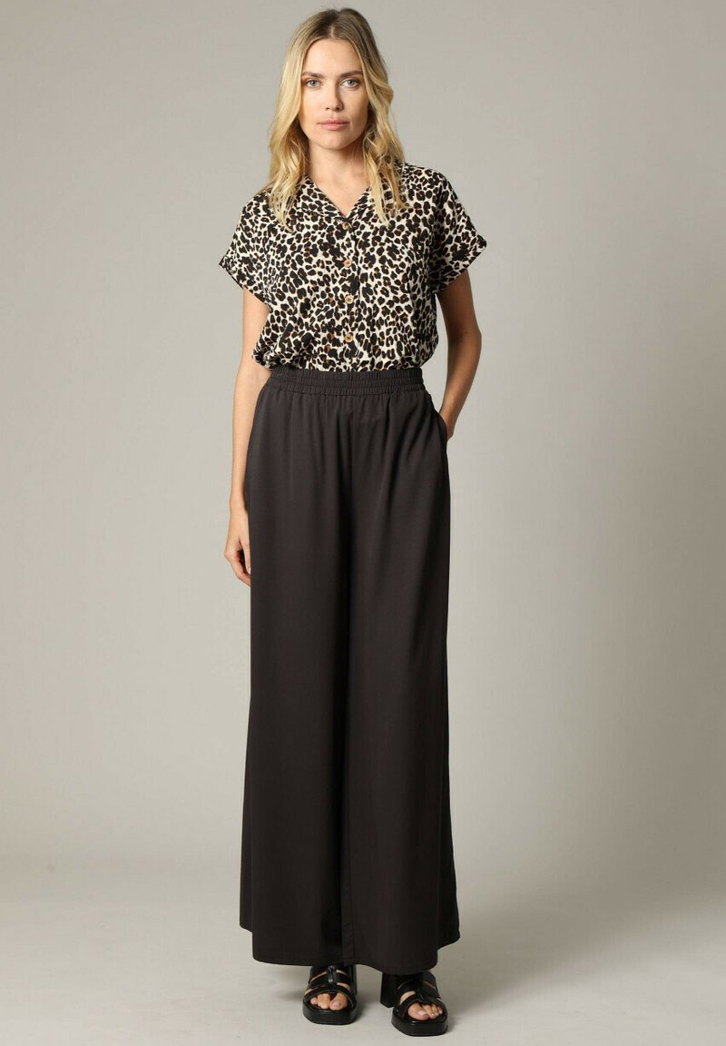 Korteblauwe blouse met luipaardpatroon, gedragen met zwarte wijde broek met elastische tailleband en platte zwarte sandalen.