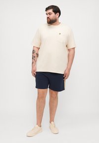 Beige bomullströja med bröstficka och gult logo, tillsammans med marinblå shorts med sidostrand, och beige sneakers.