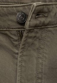 Detailný záber na olivovozelenú denimovú tkaninu, zobrazuje gombík s textom "MNG DENIM" a čiastočne viditeľný zips pod pásom.