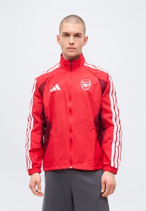 Modelo masculino vistiendo una chaqueta deportiva roja de Arsenal Adidas con rayas blancas en las mangas, posando frente a un fondo claro y liso.