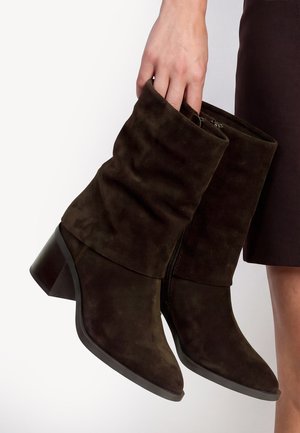 Main tenant une paire de bottes mi-mollet en daim brun foncé avec des tiges froissées et des talons carrés sur fond blanc.