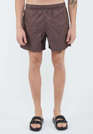 Uomo che indossa pantaloncini da bagno marroni a motivi e sandali neri con doppia fascia, mostra ginocchia e parte inferiore delle gambe con tatuaggi e piccoli segni sulla pelle.