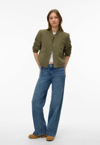 Veste bomber vert olive avec un col rond, deux poches avant, associée à un jean bleu à jambes larges et des chaussures marron. Texture de tissu lisse.